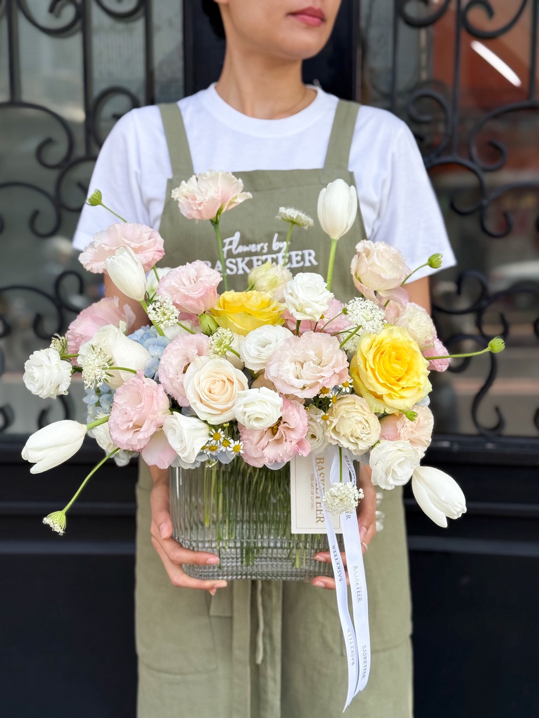 An elegant Peach Harmony Vase overflows with pastel roses, tulips, and carnations, presented before a grand black ornate door in Bangkok.