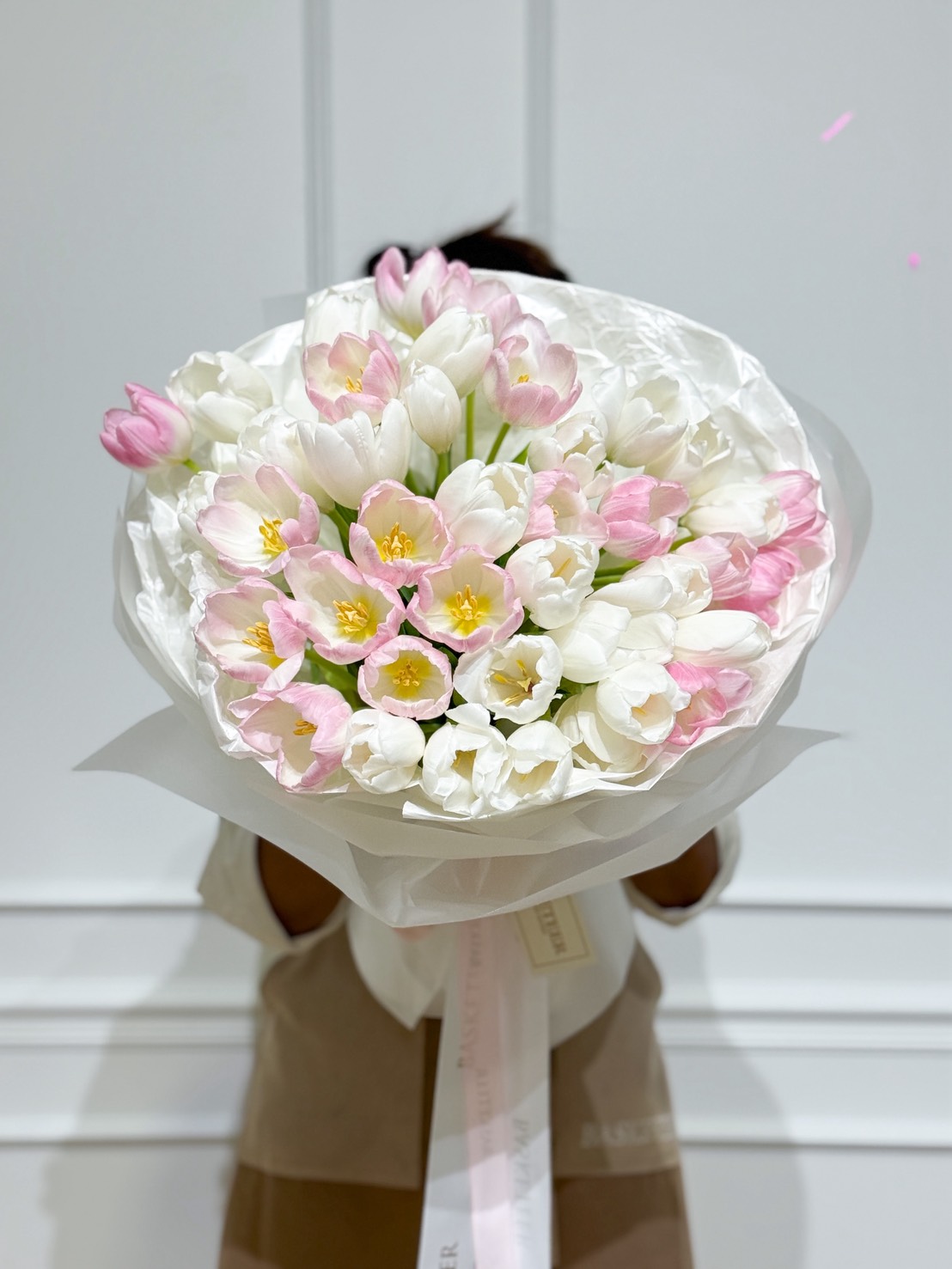 Elegant bouquet of white and blush-pink tulips wrapped in soft white paper with a Basketeer ribbon, held by florist against white wall.