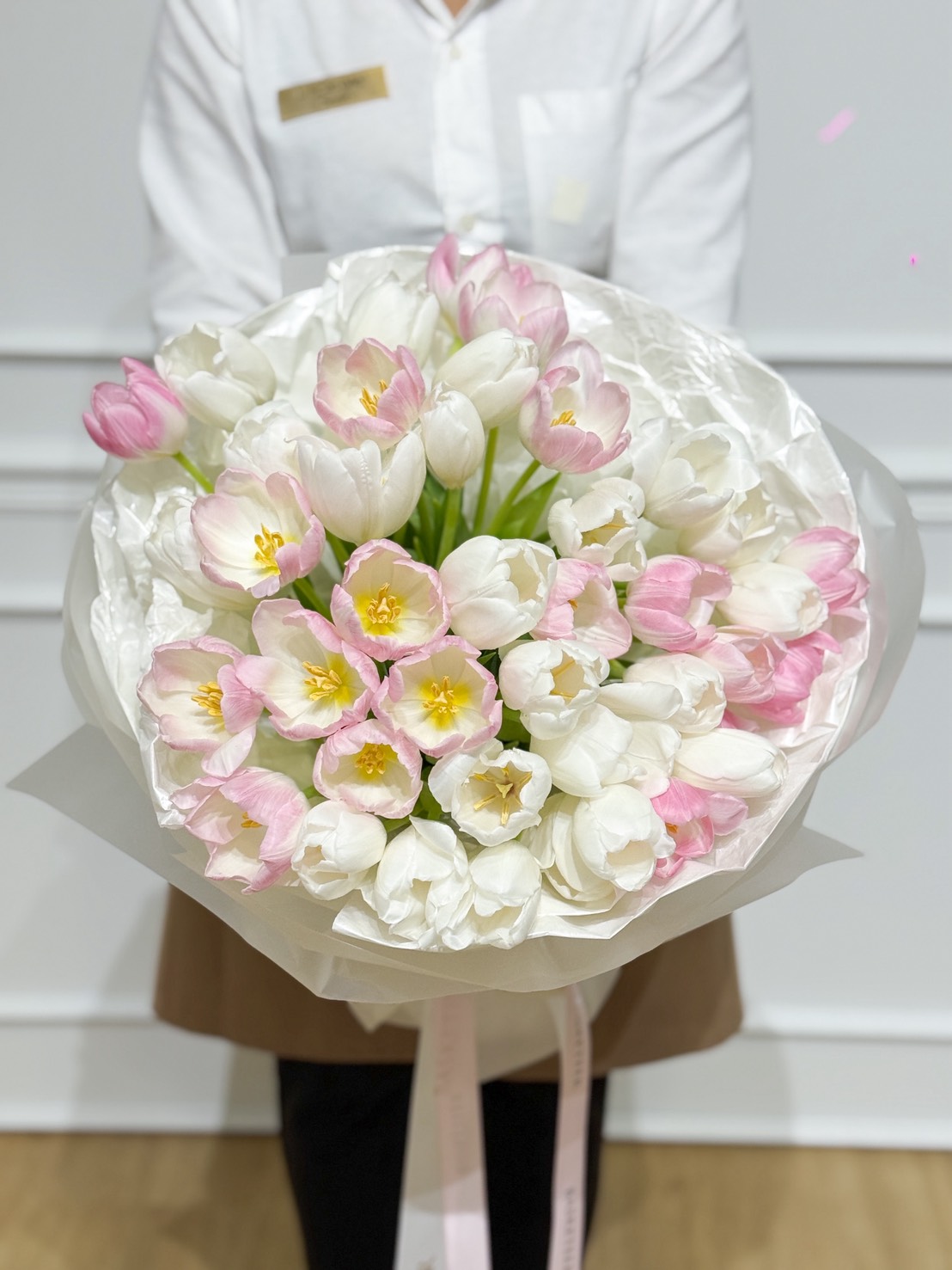 Close-up of white and blush-pink tulip bouquet wrapped in soft white paper with a Basketeer satin ribbon, held by florist in white shirt.