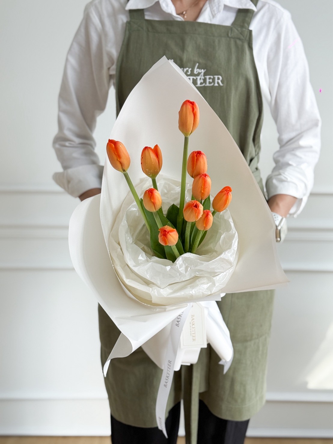 Elegant orange tulip bouquet wrapped in white paper and tied with a white Basketeer ribbon, held by a florist in a green apron.