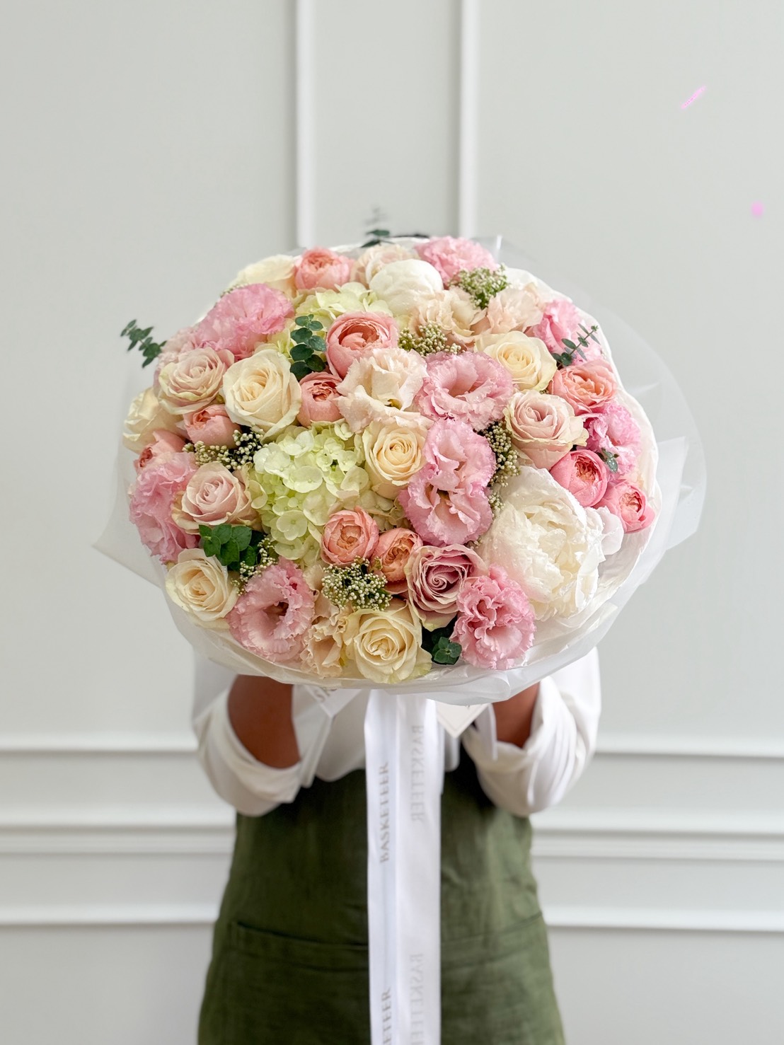 A person in a white shirt and green apron holds an Elegant Harmony Mixed Bouquet of pastel pink, white, and cream flowers wrapped in white paper, completely obscuring their face.
