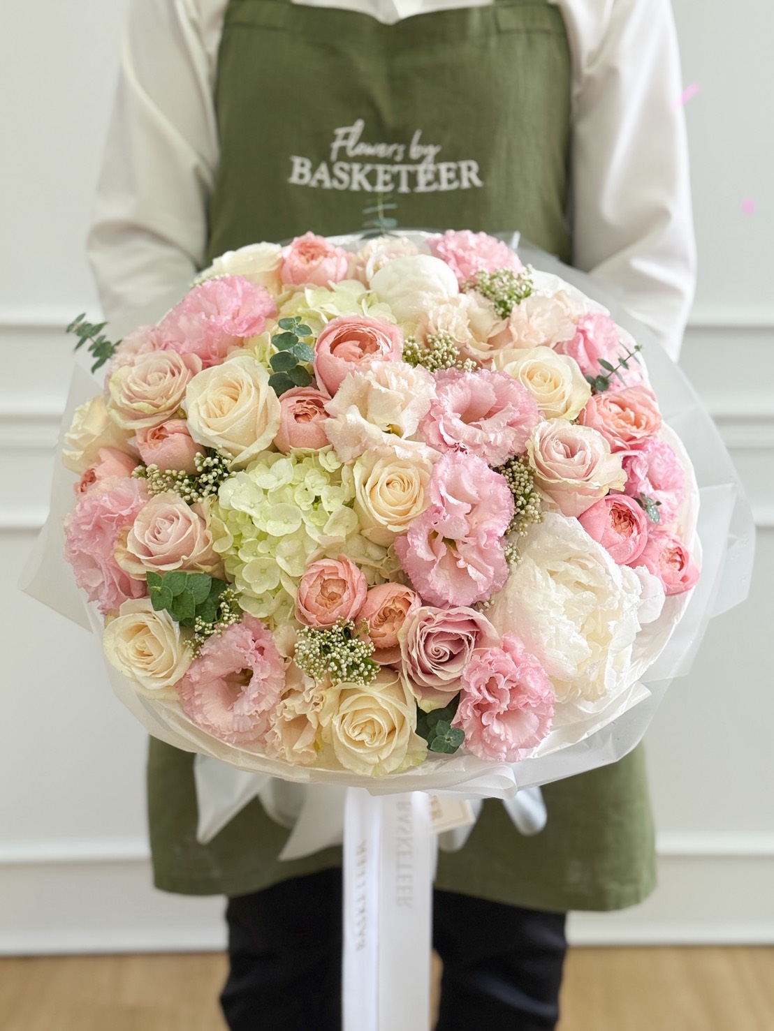 A person in a green apron holds an Elegant Harmony Mixed Bouquet—a large arrangement of pastel roses, hydrangeas, and carnations in shades of white, pink, and cream, beautifully wrapped in white paper.