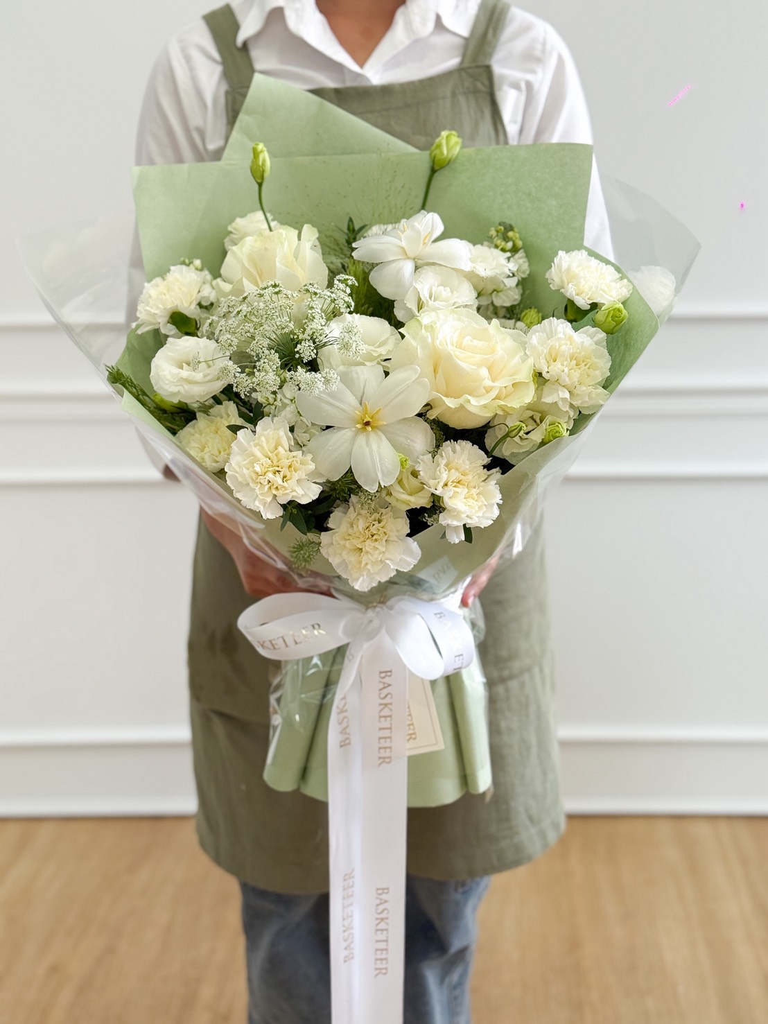 A person wearing a green apron and white shirt holds an Ivory Roses & White Chrysanthemums Bouquet, wrapped in green paper and tied with a white ribbon. The background features a white wall and wooden floor.