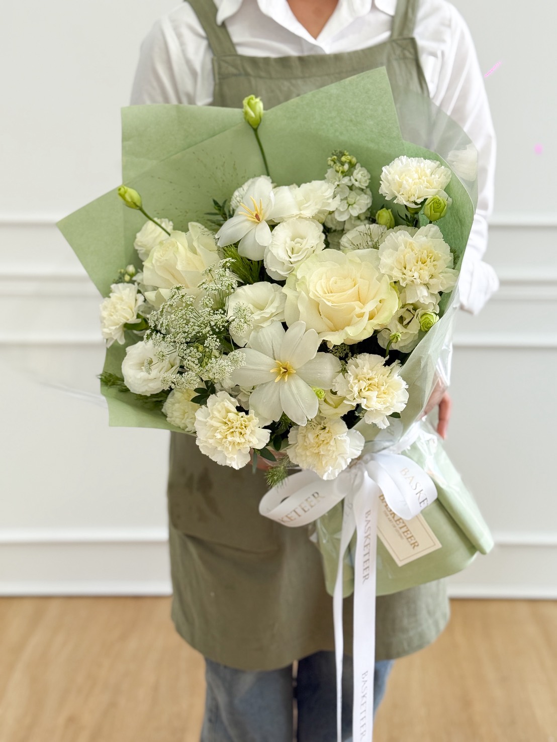 A person wearing a green apron holds an Ivory Roses & White Chrysanthemums Bouquet, wrapped in green paper and tied with a white ribbon, standing indoors on a wooden floor.