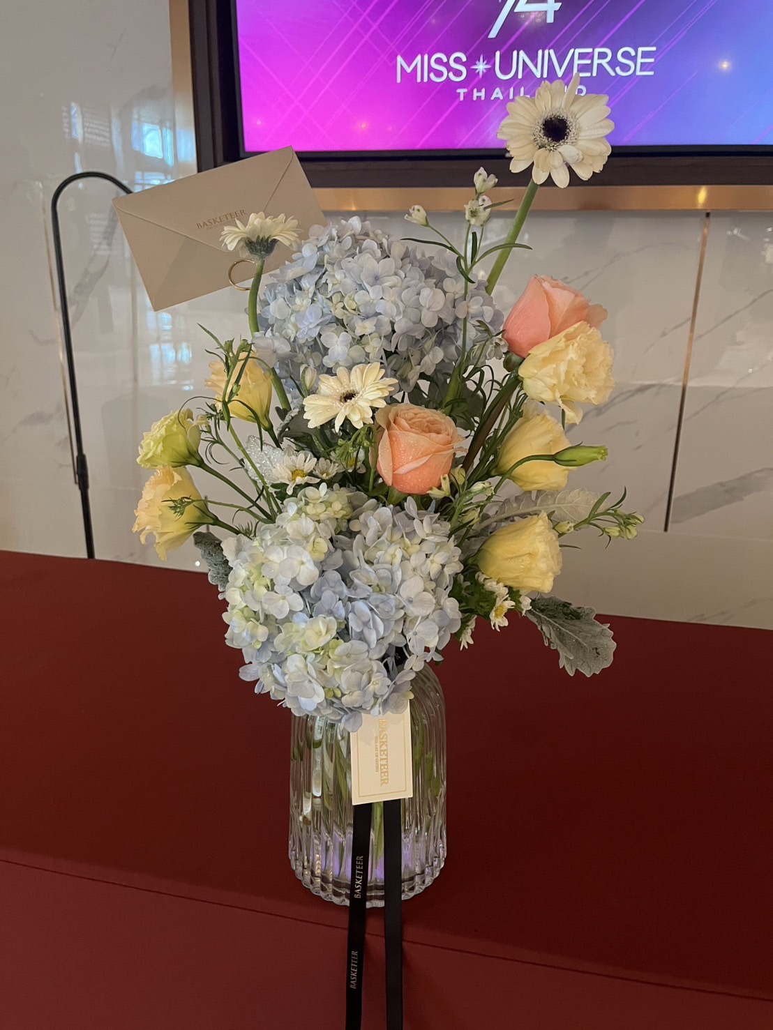 A glass Radiant Harmony Vase Arrangement holds blue hydrangeas, peach roses, white daisies, and yellow blooms. A card is tucked among the flowers. In the background, a TV displays “Miss Universe Thailand.”.