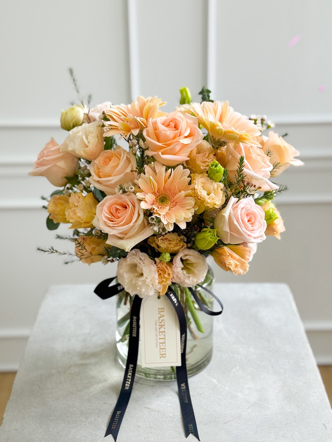 The Peach Elegance Vase features a bouquet of peach roses, gerbera daisies, and carnations with lush greenery. Tied with a black 