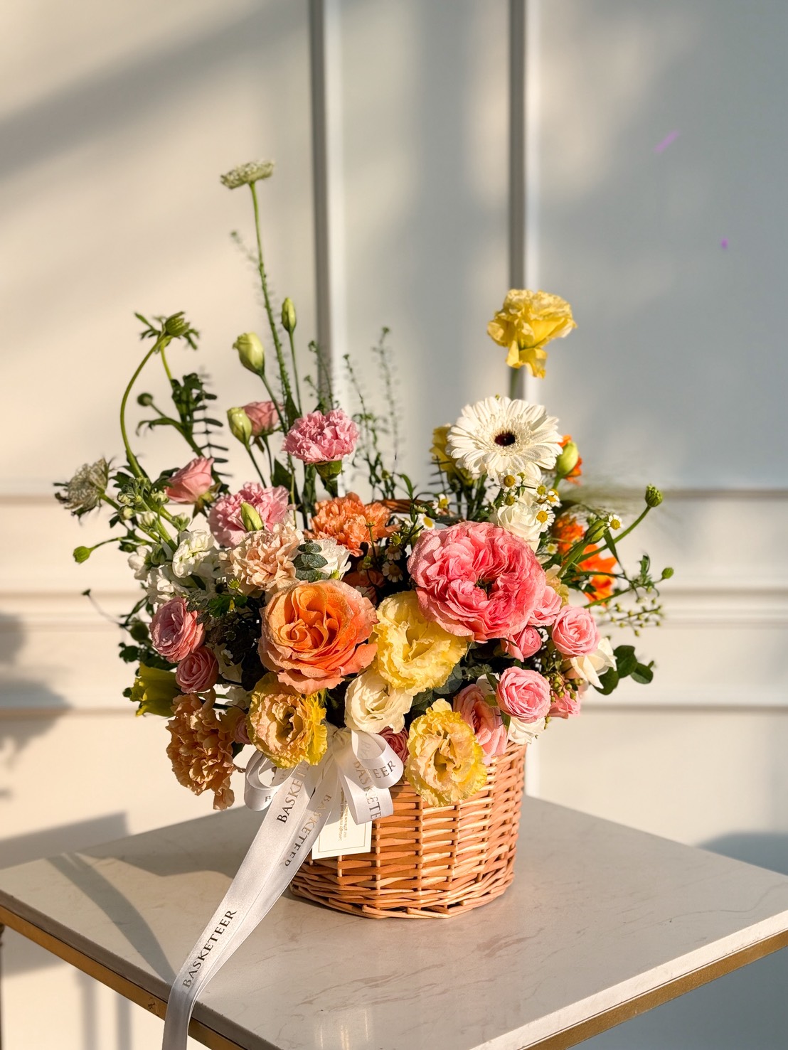 The Radiant Harmony Flower Basket, filled with vibrant pink, yellow, orange, and white blooms, sits on a white table against a softly lit wall. A delicate white ribbon is tied round the wicker basket.