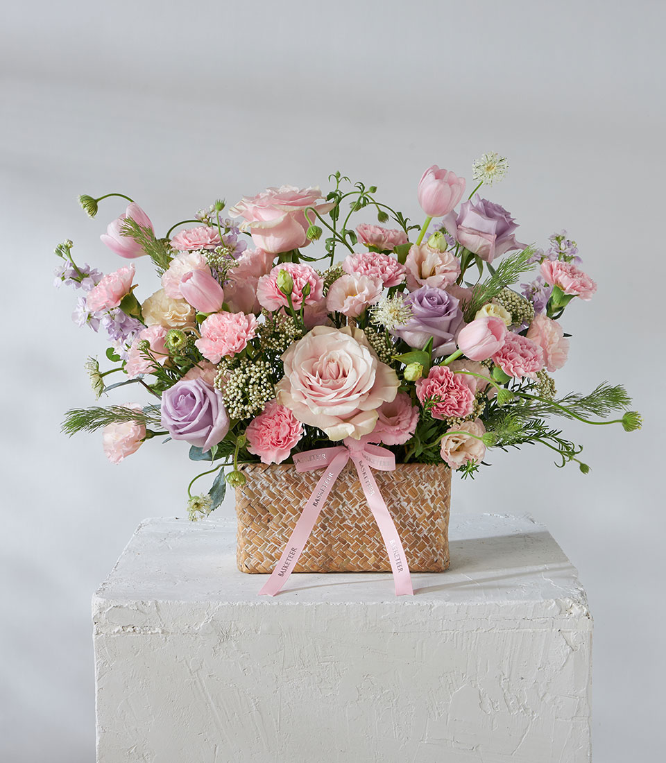 A Lavender Bloom Flower Basket filled with pink and lavender roses, carnations, tulips, and greenery sits against a plain light background, adorned with a charming pink ribbon tied round the woven basket.