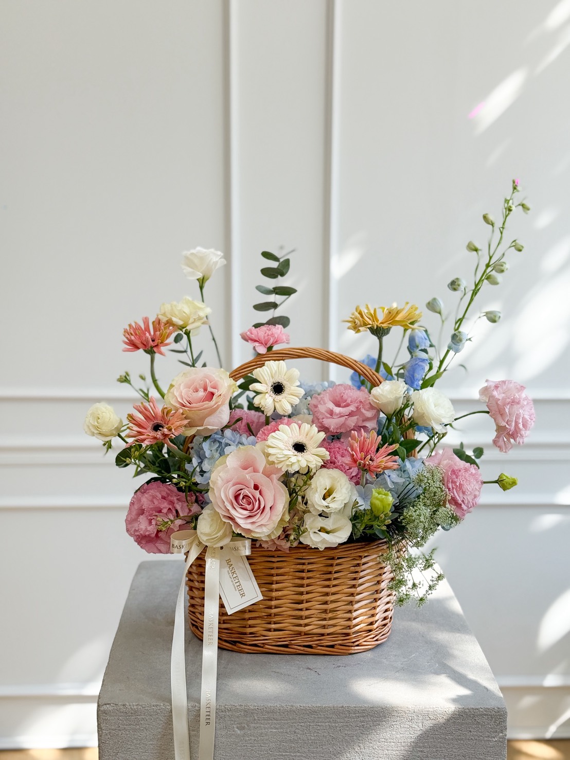 A Pastel Bloom Flower Basket filled with assorted pastel flowers, including roses, daisies, and carnations, sits on a grey pedestal against a white panelled wall. A ribbon and small card are attached to the basket handle.