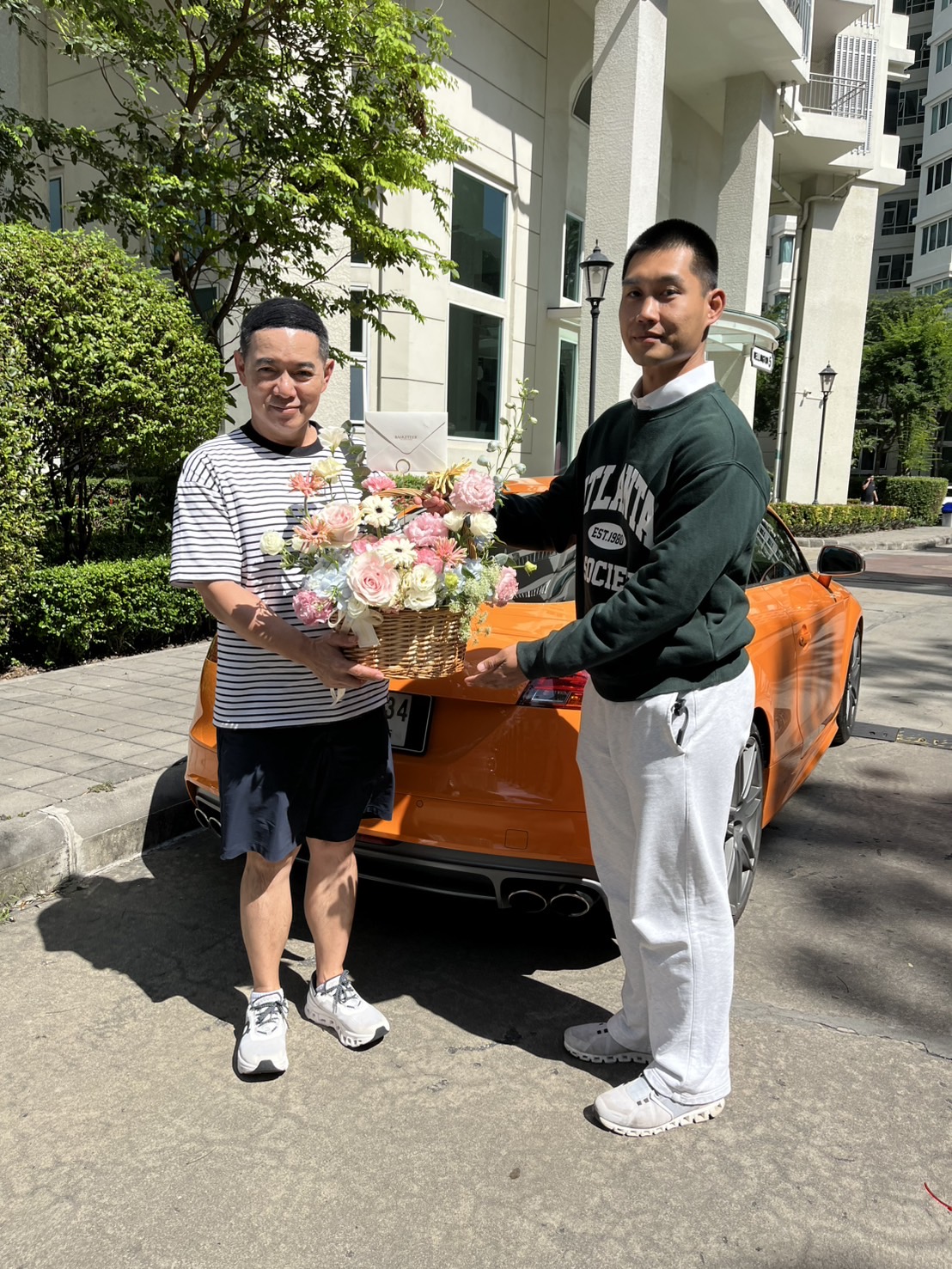 Two men stand outdoors in front of an orange sports car, smiling as they hold a large Pastel Bloom Flower Basket together, with greenery and a white building in the background.