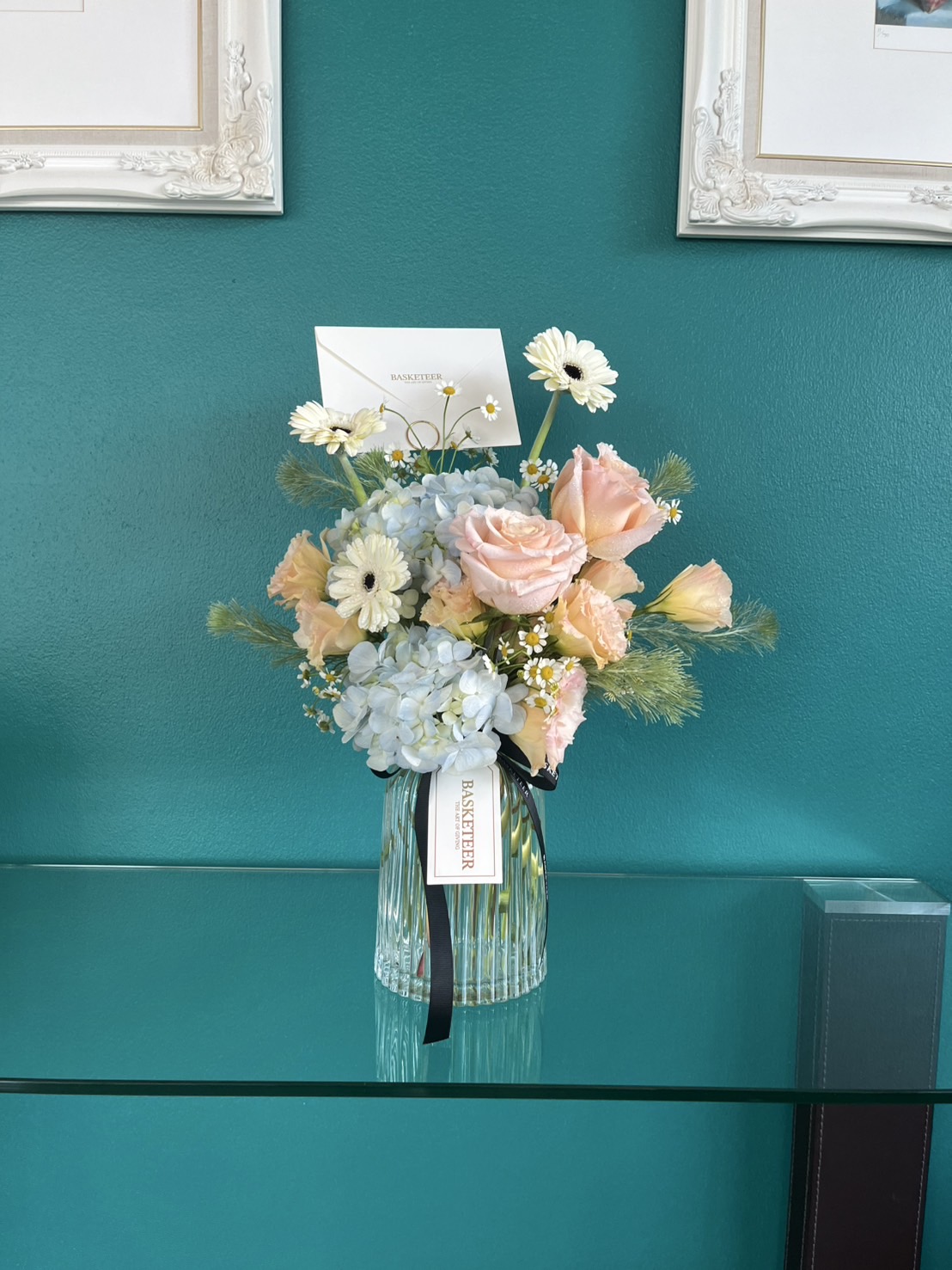 A Radiant Harmony Vase Arrangement holds a bouquet of pastel flowers—pale pink roses, blue hydrangeas, white daisies, and greenery. A small card is tucked among the blooms on a glass shelf against a teal wall.