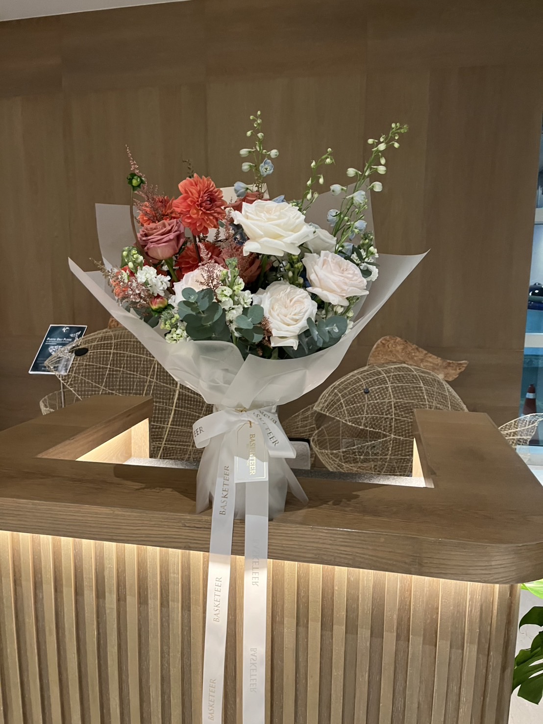 An Ivory Rose & Blue Delphinium Bouquet, featuring white roses and red blooms, is wrapped in white paper and tied with a white ribbon. The elegant bouquet is displayed on a wooden reception counter with modern chairs in the background.