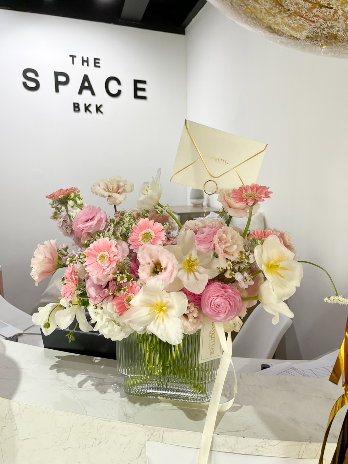 A Graceful Symphony Vase filled with pink and white flowers sits on a marble counter. Behind it, a white wall displays the words 