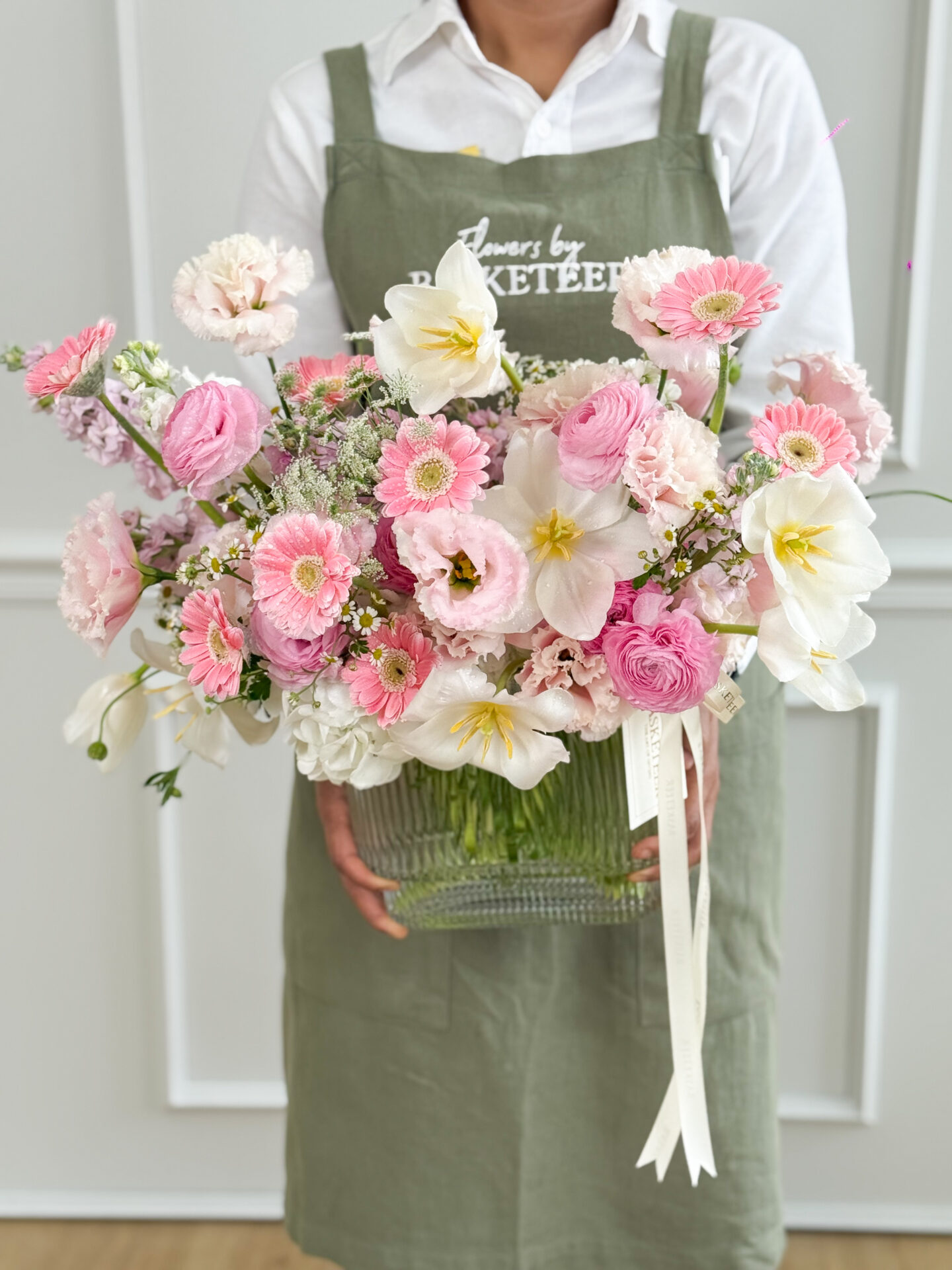 A person in a green apron holds the Graceful Symphony Vase, filled with a variety of pink and white flowers, including roses and daisies, against a light-coloured indoor background.