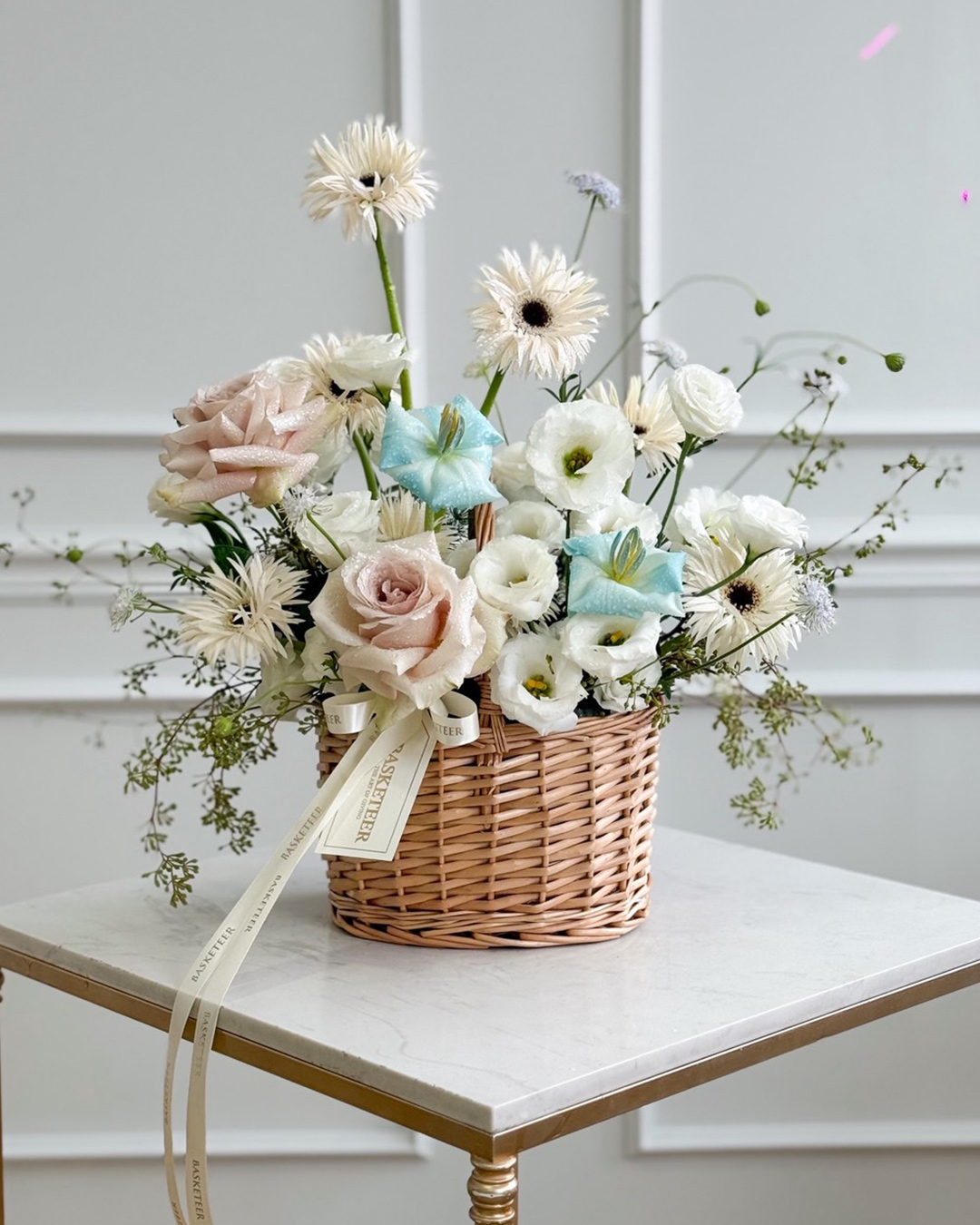 Luxury pastel flower basket with roses, white lisianthus, blue flowers, and white gerberas in natural woven basket.