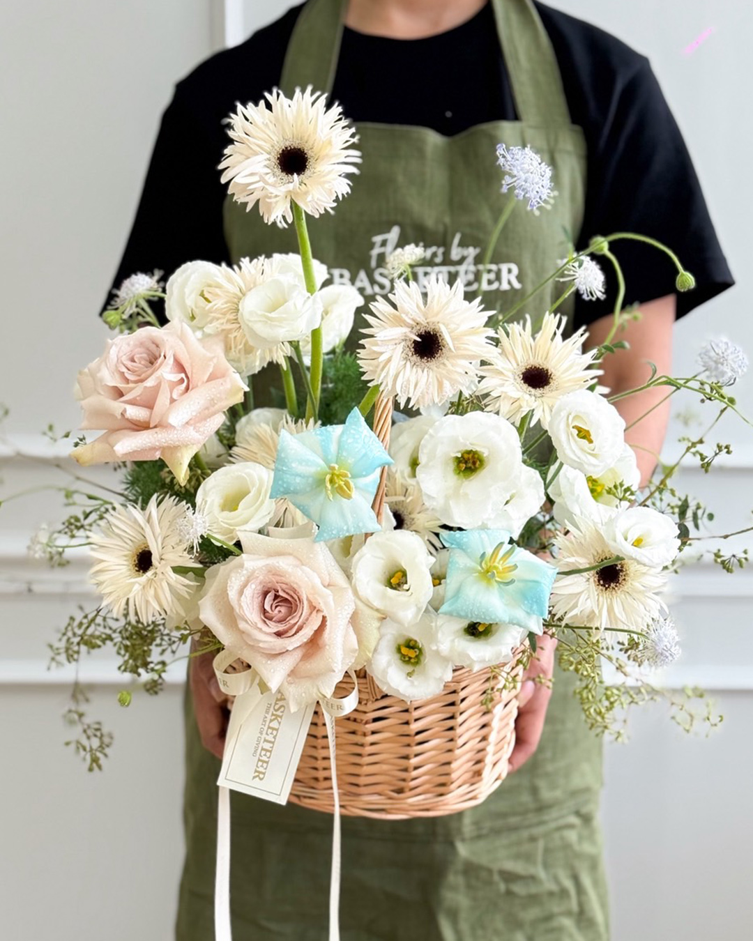 A person in a green apron holds the Blue Serenity Flower Basket, filled with white and pale pink roses, daisies, and assorted flowers, accented with blue and white ribbons and a tag.