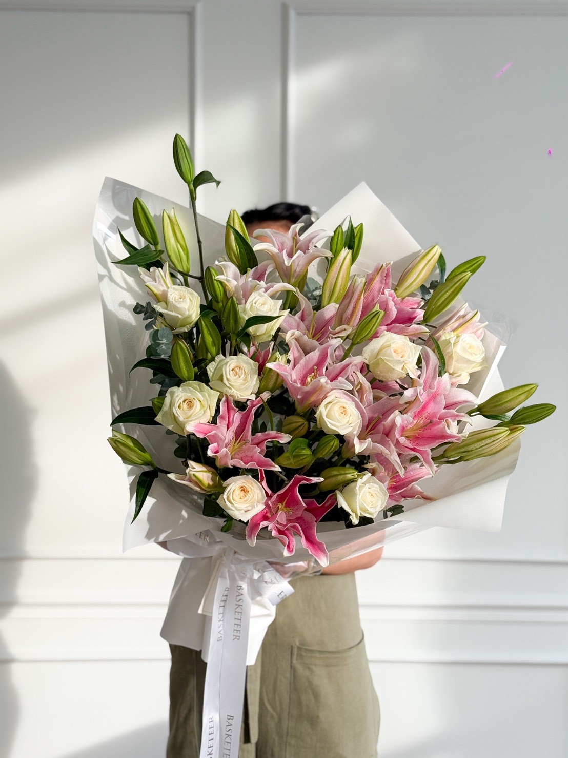 A person holds a Grand Pink Oriental Lily Bouquet with white roses, wrapped in white paper and tied with a white ribbon. The background features a white wall adorned with decorative moulding.
