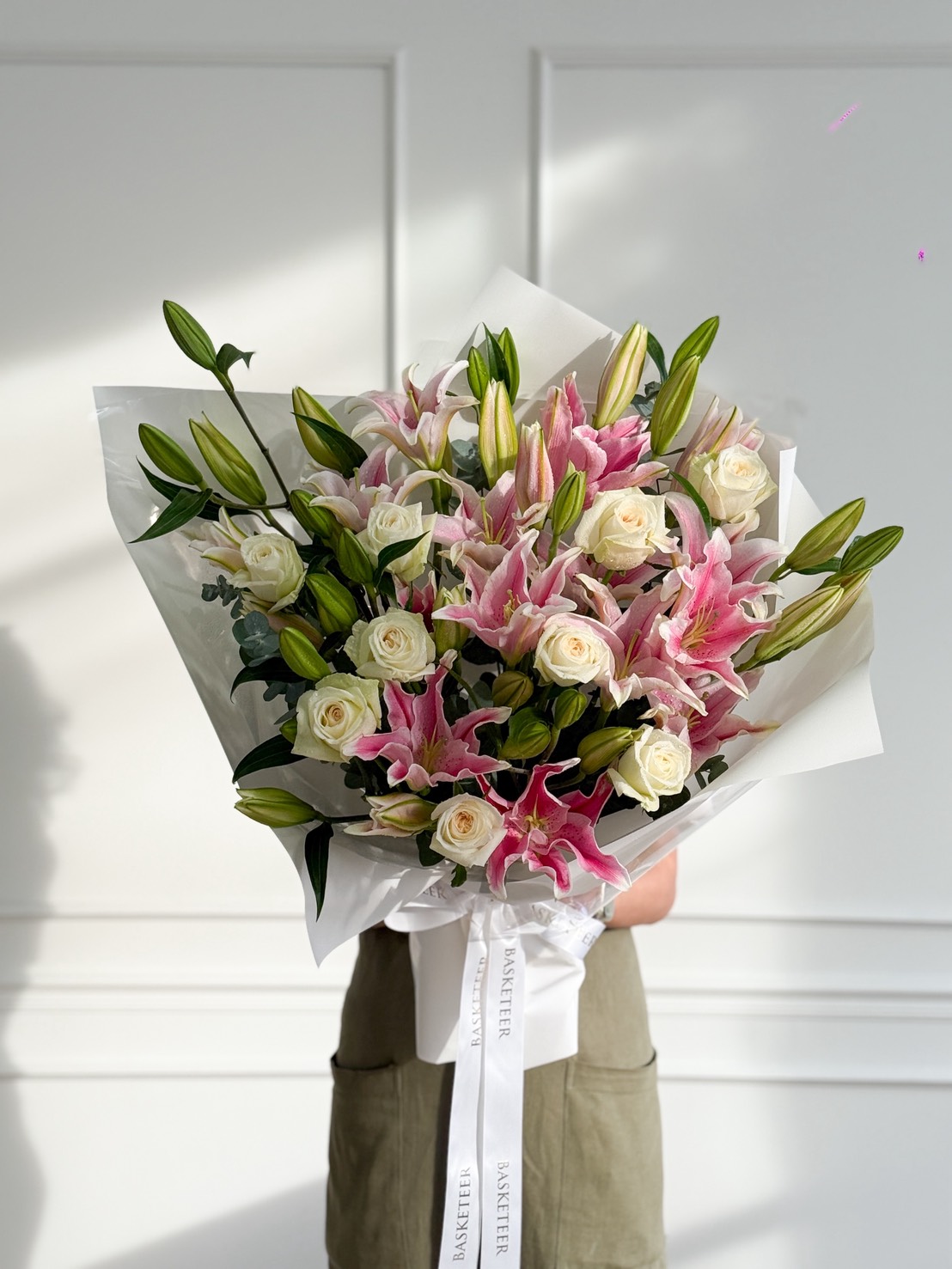 A person holds a Grand Pink Oriental Lily Bouquet of white roses and pink lilies wrapped in white paper with a white ribbon, standing against a white wall. The large bouquet completely obscures the person's face.