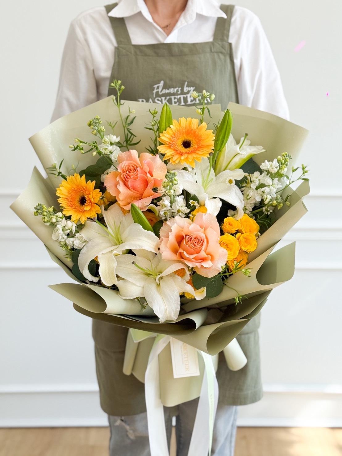 A person wearing an apron holds a stunning White Lilies & Gerbera Bouquet with peach roses, yellow gerbera daisies, and assorted greenery, all elegantly wrapped in pale green paper and tied with a white ribbon.