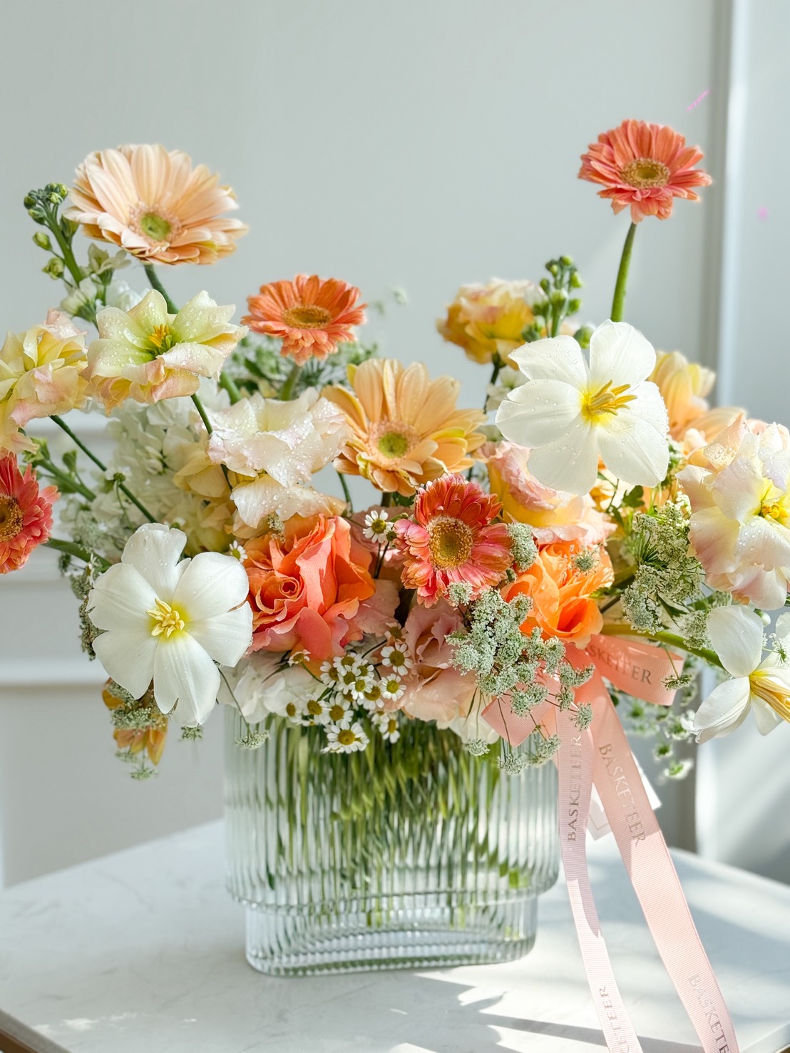 The Peach Harmony Vase Arrangement features a glass vase holding a vibrant bouquet of orange, peach, white, and yellow flowers with greenery. Pink ribbons adorn the vase, as sunlight highlights the display against a soft background.