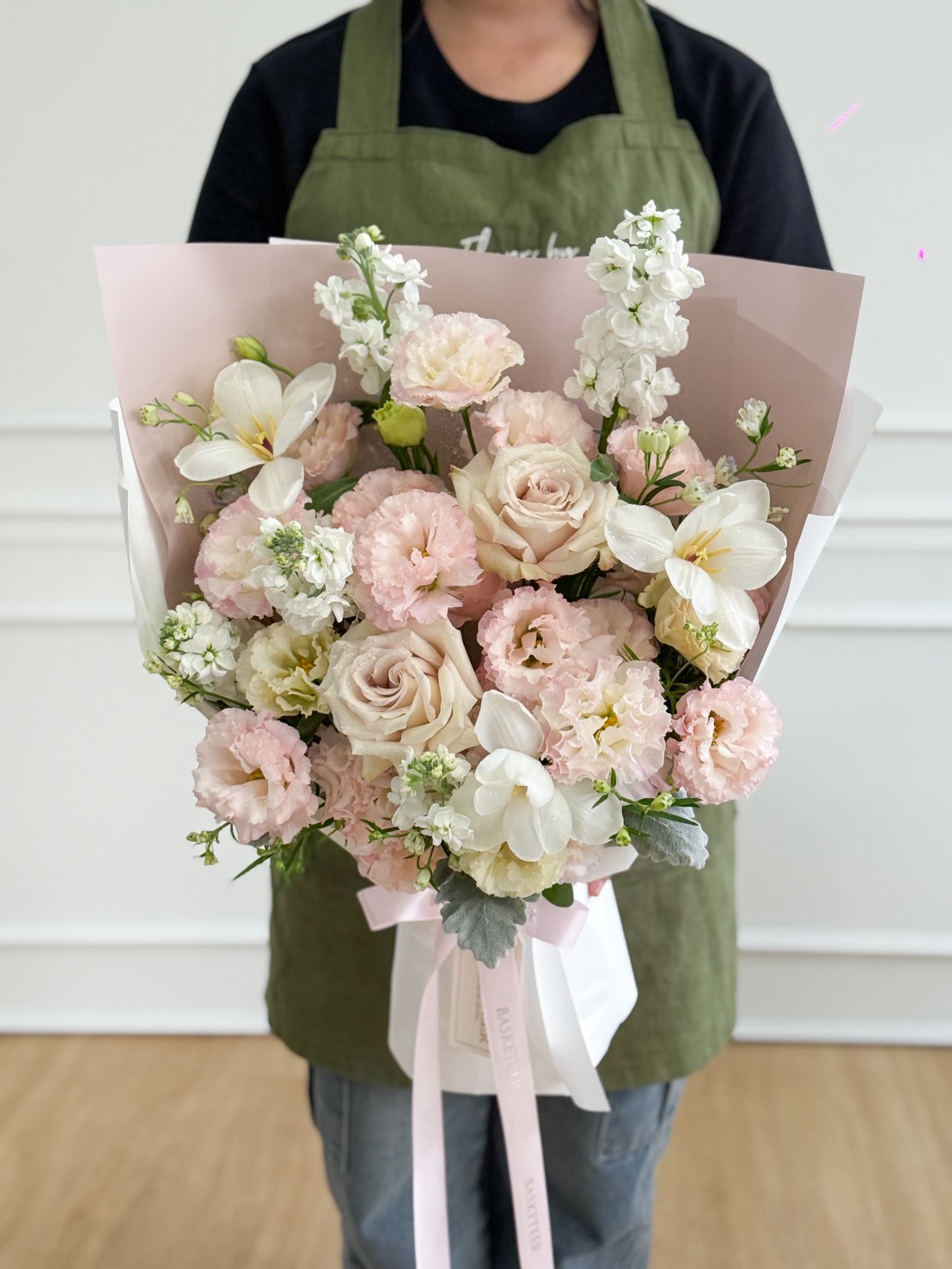 A person in a green apron holds an Ivory & Blush Roses with White Freesia Pastel Bouquet, featuring pale pink and white flowers like roses and lilies, all wrapped in pastel pink paper with a white ribbon.