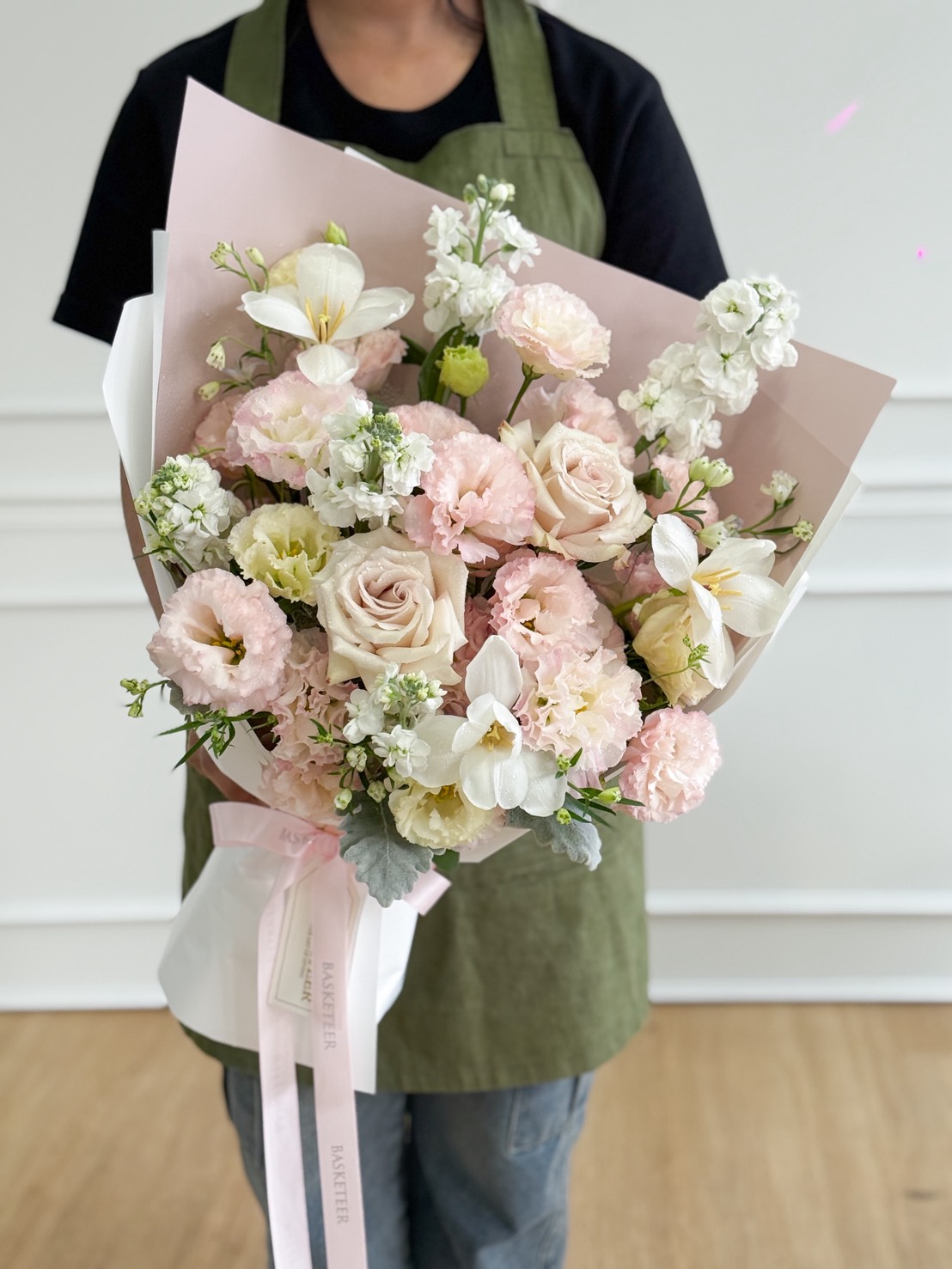 A person in a green apron holds an Ivory & Blush Roses with White Freesia Pastel Bouquet, featuring pastel pink and white flowers, including roses and lilies, wrapped in pale pink paper and tied with a white ribbon.