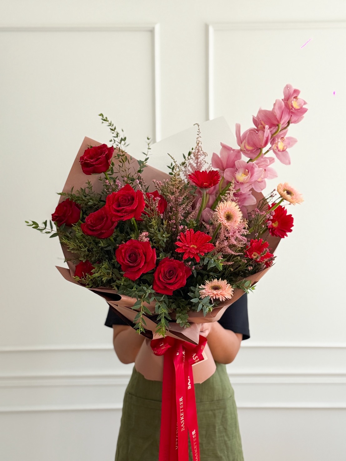 A person in a dark shirt and green skirt holds the Eternal Grace Bouquet—red roses, pink orchids, and more—wrapped in paper and tied with a red ribbon, all set against a white wall background.