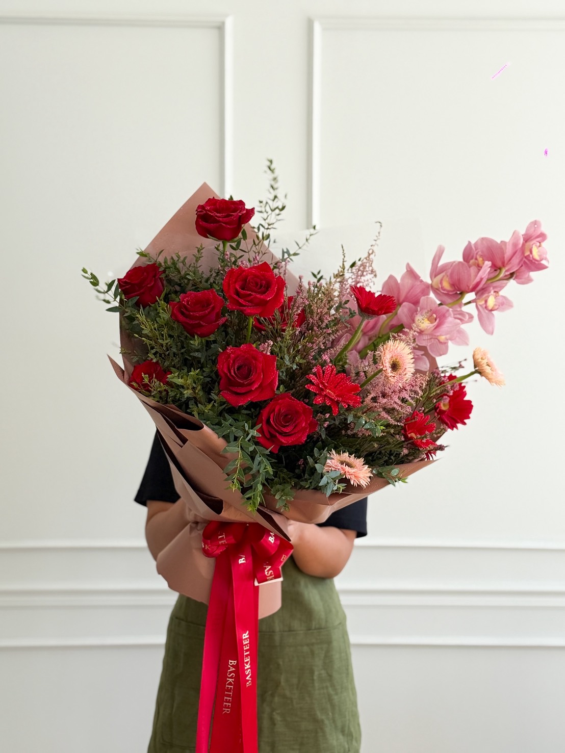 A person in a black top and green skirt holds the Eternal Grace Bouquet—a large arrangement of red roses, pink orchids, and various flowers wrapped in paper with a red ribbon—standing against a white wall.