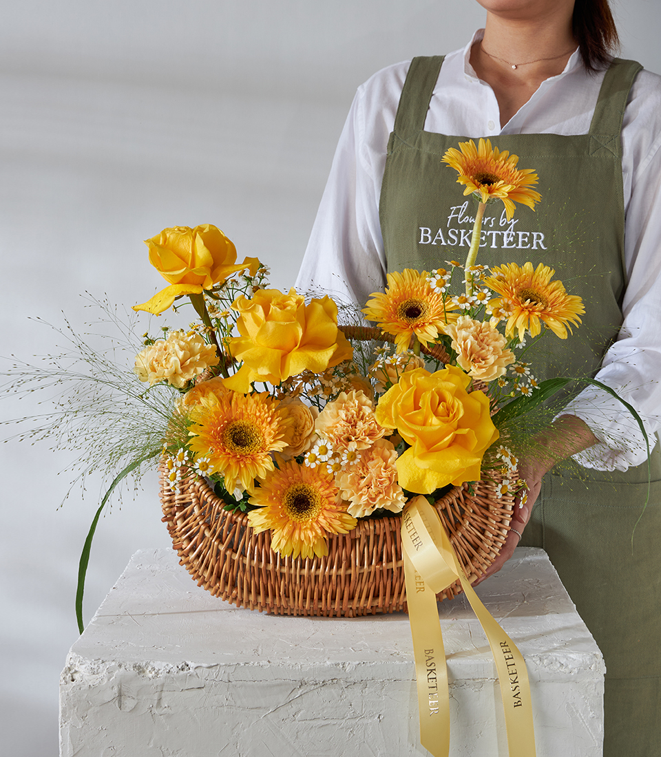 A person in a green apron holds the Golden Radiance Basket, filled with yellow roses, gerbera daisies, carnations, lush greenery, and a yellow ribbon, all beautifully arranged on a white pedestal.