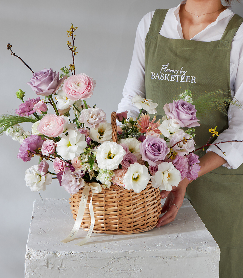 A person in a green apron arranges a Lavender Grace Flower Basket filled with pastel flowers, including white, pink, and lavender blooms, on a white textured plinth. The apron reads 
