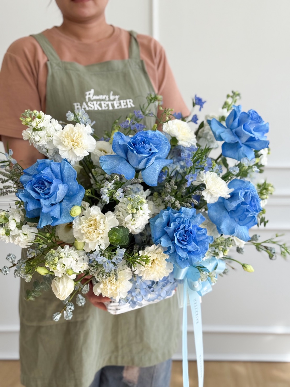 A person in an apron holds the Whisper Blue Elegance Basket, a large bouquet featuring blue roses, white carnations, and assorted greenery, beautifully arranged in a white basket with a light blue ribbon.