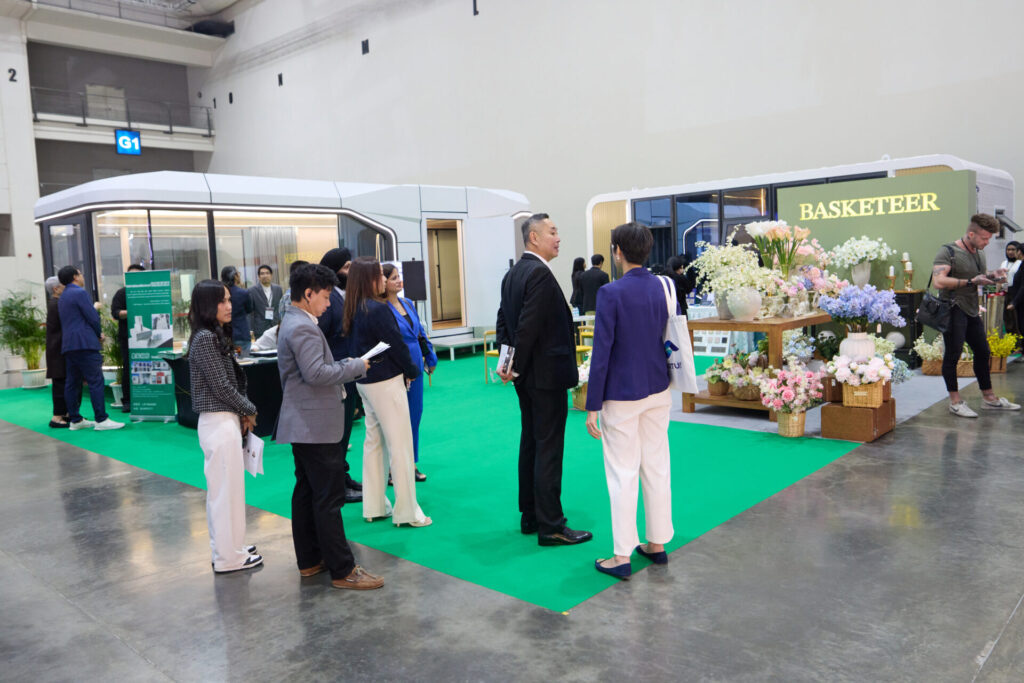 People in business attire stand and converse near a green-carpeted exhibition stand labelled "BASKETEER," featuring baskets and floral displays. Modern structures and promotional materials are visible in the background.