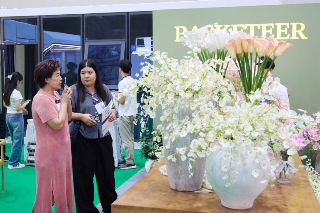 Two women stand talking near a display of large flower arrangements in white vases at an indoor event. Other people are in the background near a green wall with the word "PAKETTEER" partially visible.