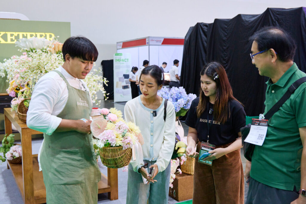 A florist in an apron demonstrates flower arranging to three people at an indoor event. They are gathered around a display of flower baskets, and there are informational stands and signs in the background.