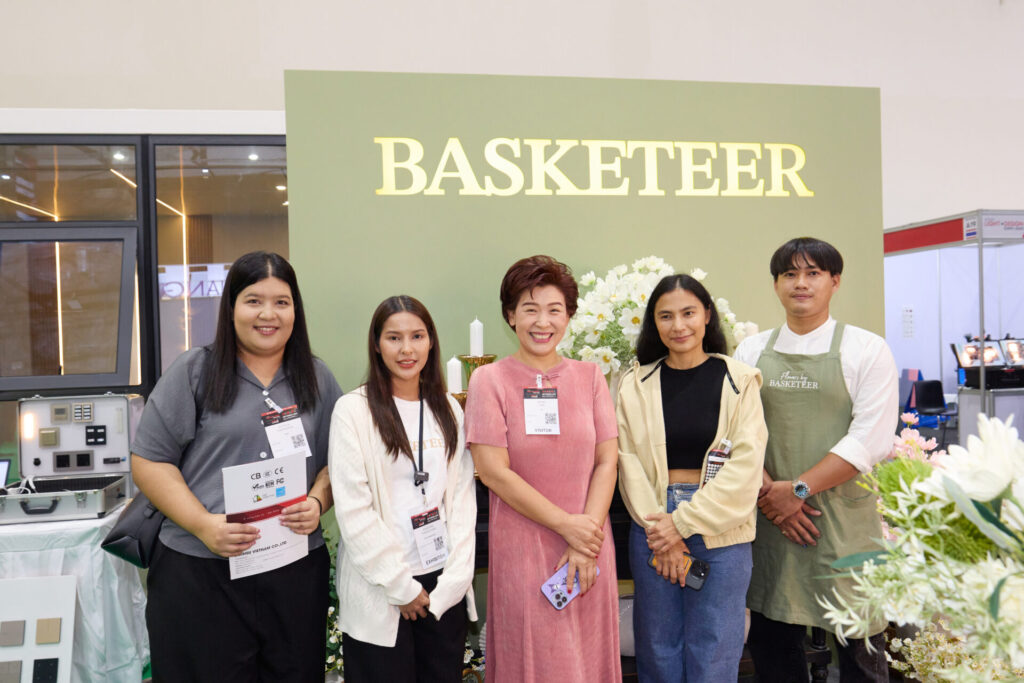 Five people stand smiling in front of a green backdrop with the word “BASKETEER” in large letters. They are indoors and surrounded by flowers and event stands.