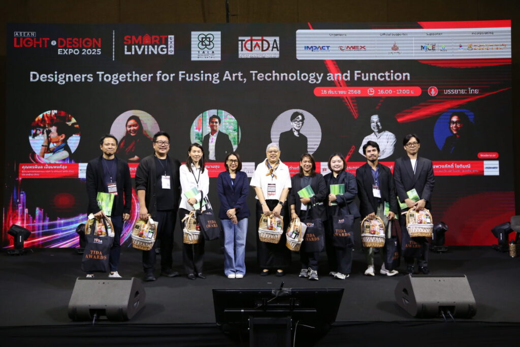 A group of nine people stands on a stage holding gift bags, posing for a photo at a design expo event. Behind them, a large screen displays event details and portraits of speakers.