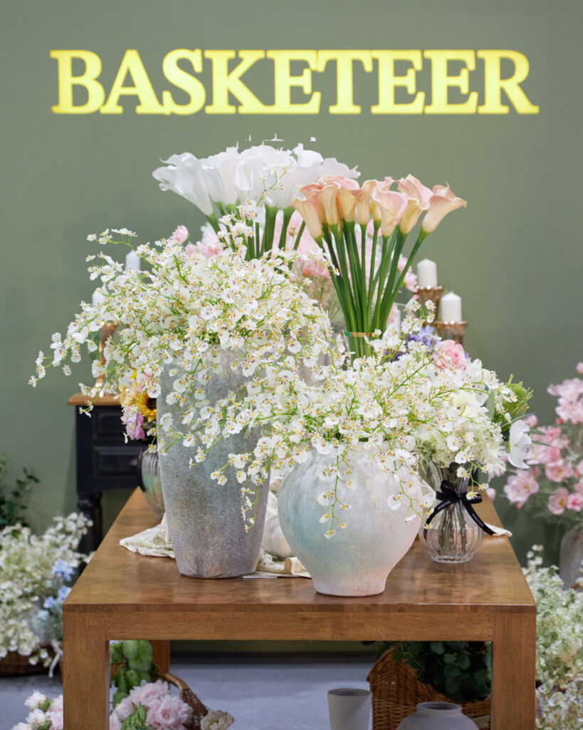 A wooden table displays large vases filled with white and pale pink flowers, set against a green wall with the word "BASKETEER" in bold yellow letters. More flower arrangements decorate the background.