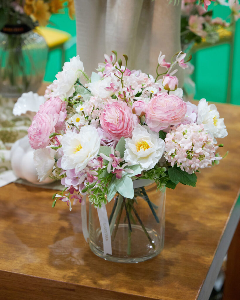 A clear glass vase filled with a bouquet of pink and white flowers, including roses and peonies, sits on a wooden table. The background features soft, blurred colours and decorative elements.