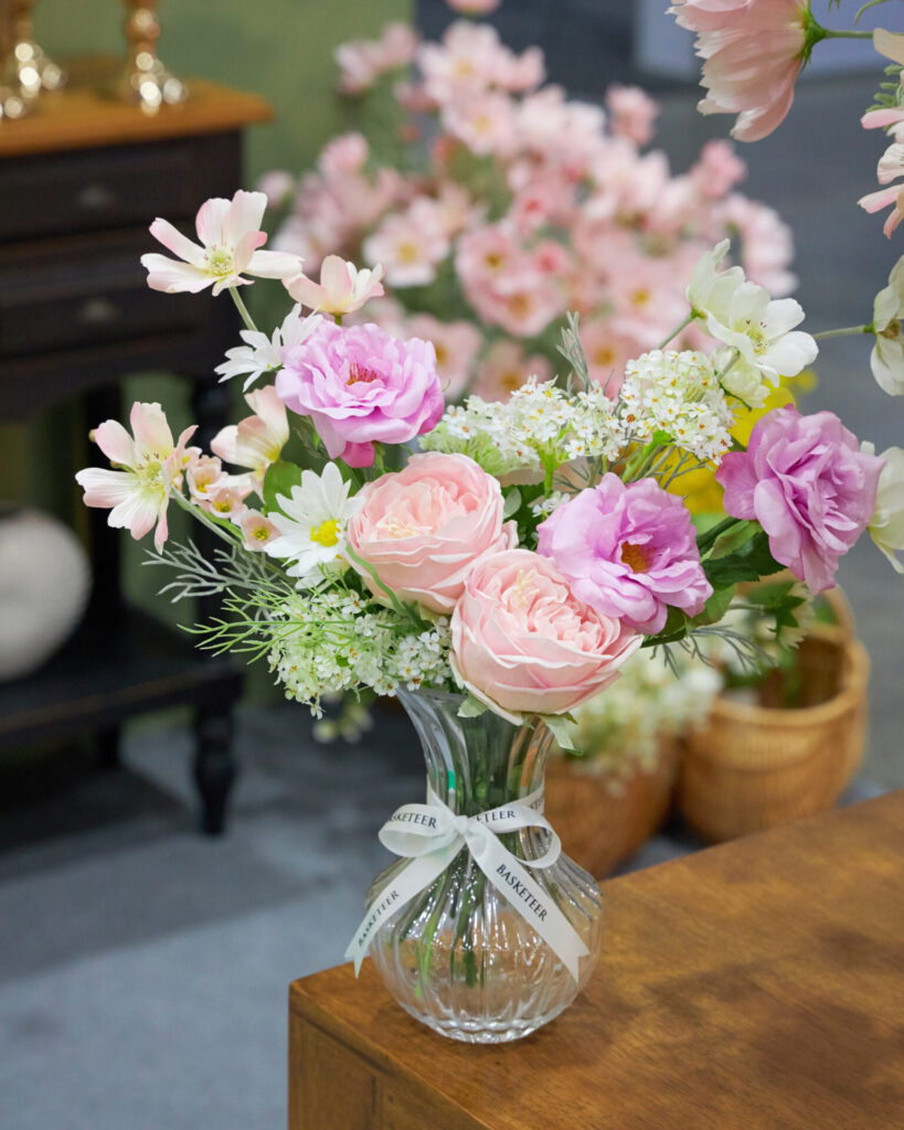 A clear glass vase filled with pink roses, light pink and white flowers, and greenery sits on a wooden table. The vase has a ribbon tied around it. Blurred pink flowers and baskets are in the background.