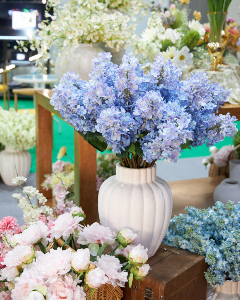 A white ceramic vase filled with blue hydrangeas is displayed on a wooden table, surrounded by various arrangements of pink, white, and blue flowers in a bright indoor setting.