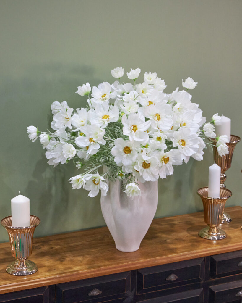A white ceramic vase filled with white flowers sits on a wooden table, flanked by two glass candle holders with white candles, against a green wall background.