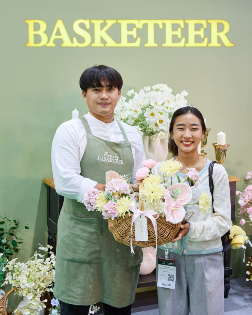 A man in a green apron and a woman holding a basket of flowers stand smiling in front of a "BASKETEER" sign with flower arrangements and candles in the background.