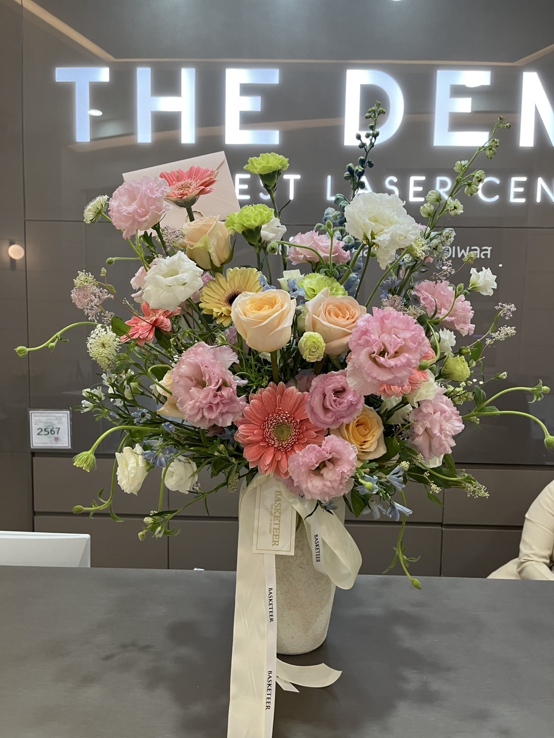 A bouquet of Juliet Roses & pastel garden flowers arranged in a ceramic vase with a white ribbon, placed on a reception counter in front of a sign that reads 