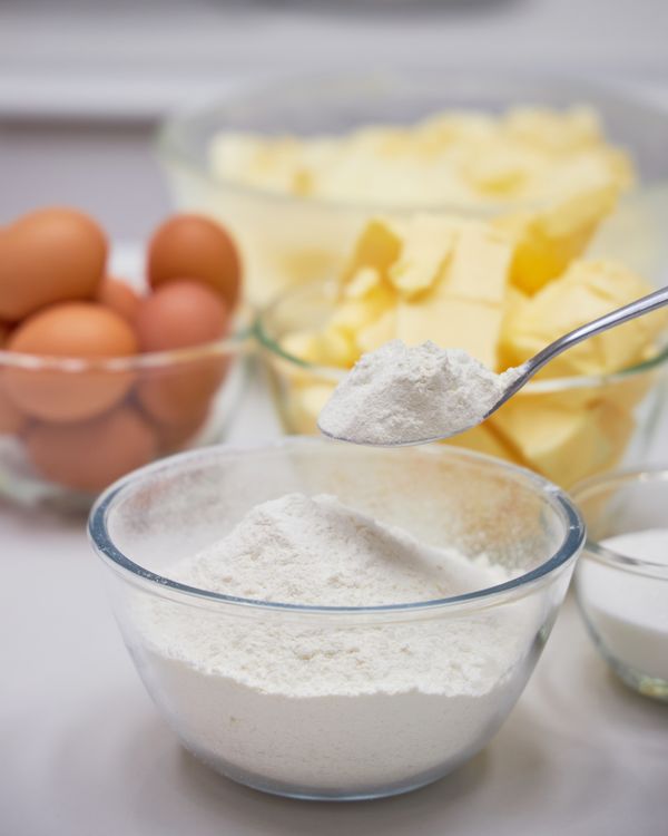 A glass bowl of flour with a spoon poised above, alongside eggs and butter, ready for a classic British Charlotte cake in Bangkok.