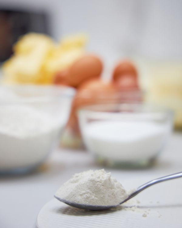 A spoonful of flour held above classic cake ingredients—sugar, eggs, and butter—ready for a Charlotte recipe in a Bangkok kitchen.
