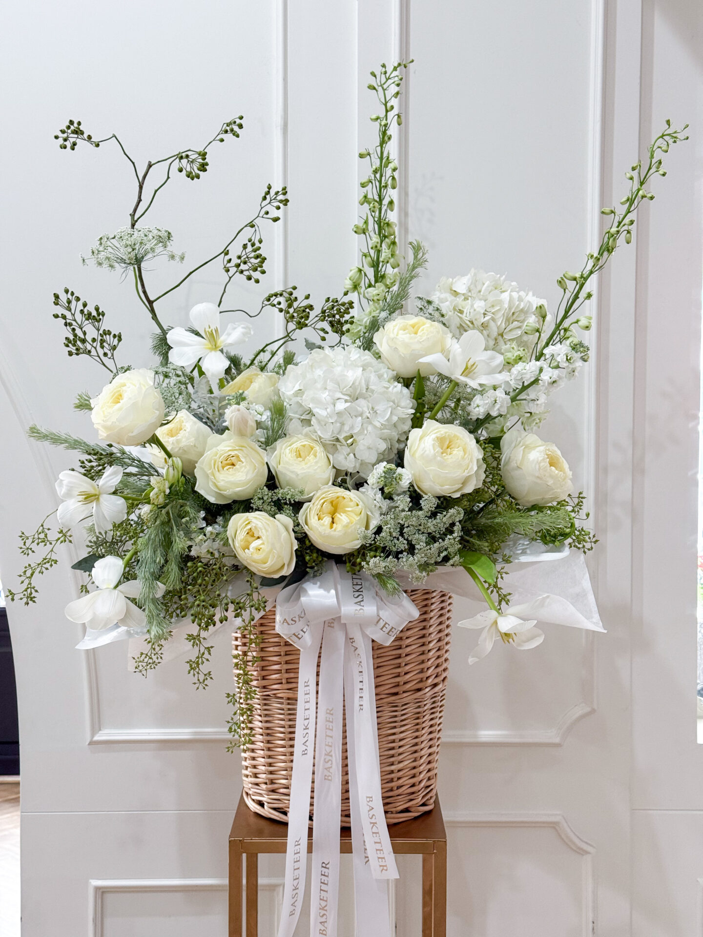 A white flower arrangement featuring roses, hydrangeas, lilies, and delicate greenery arranged in a woven basket with long white ribbons, placed against a white paneled background.