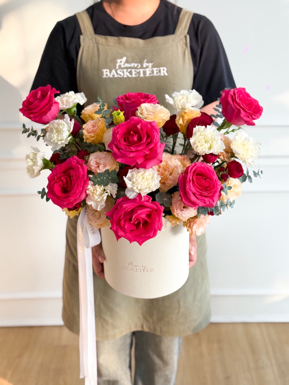 A person wearing a green apron holds a Full Monty Red Roses & White Lisianthus Flower Box, its vibrant red roses and lisianthus mixed with pale carnations and greenery, arranged in a lush bouquet. 