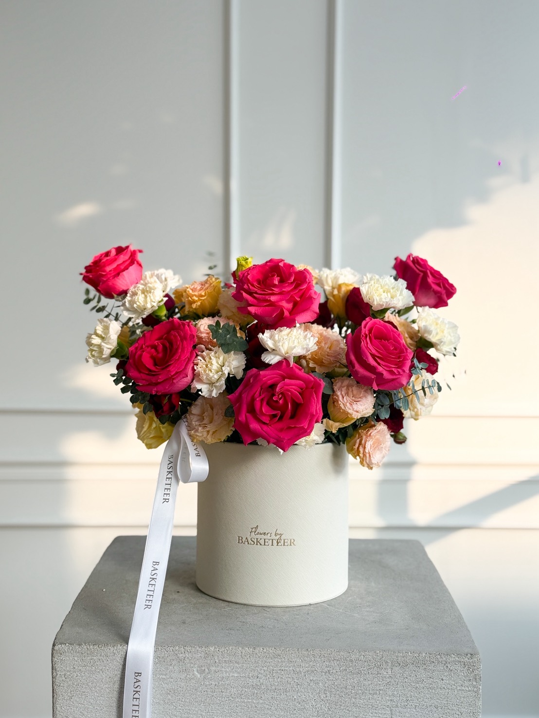 A white box filled with vibrant Full Monty Red Roses & White Lisianthus Flower Box sits on a grey pedestal. The box, tied with a white ribbon and labelled “BASKETEER,” is gently illuminated as sunlight casts soft shadows behind.