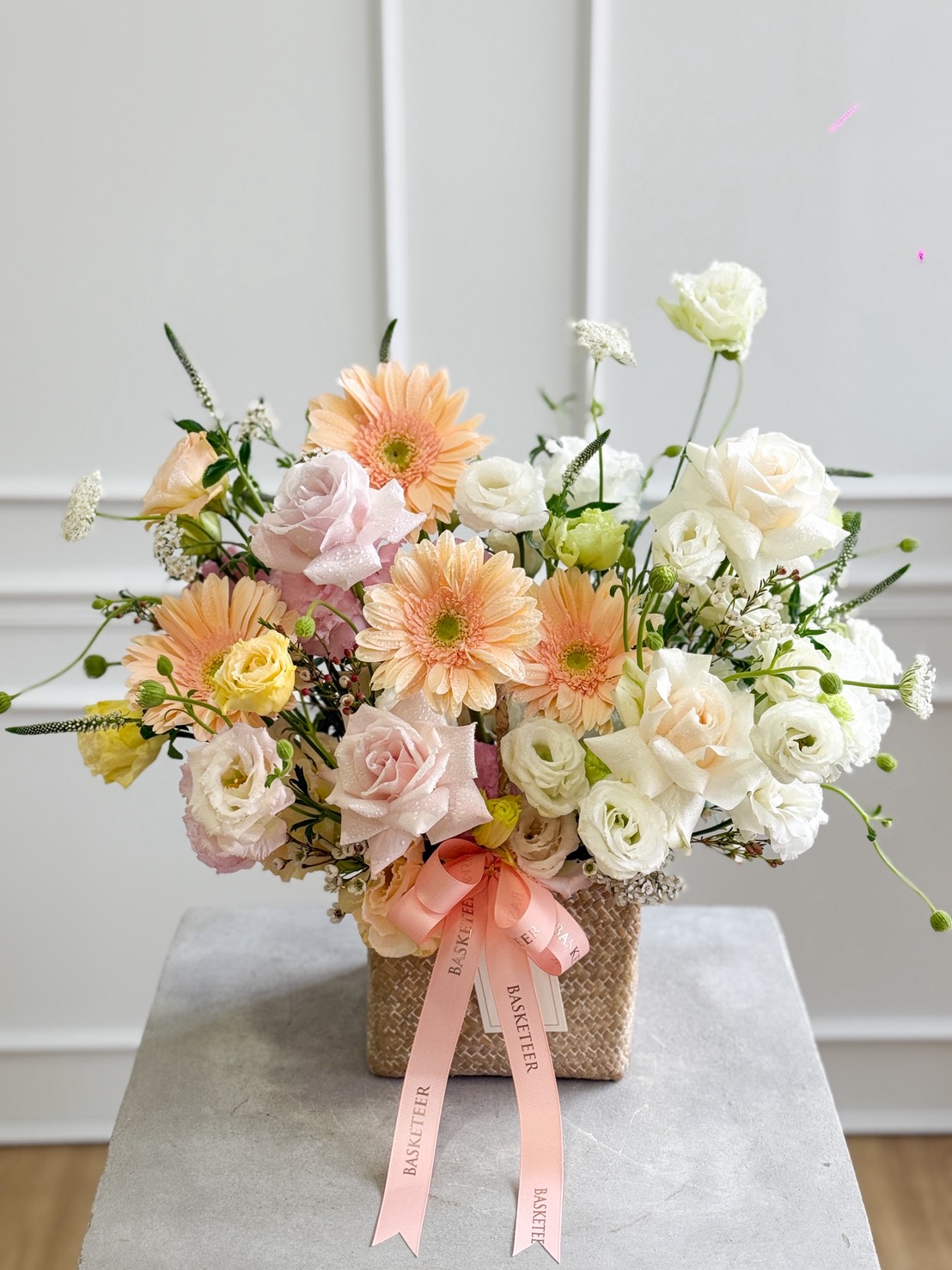 A woven basket filled with pastel flowers, including Peach Gerberas, White O’Hara & Pink Mondial Roses Pastel Basket blooms, sits on a grey surface. A pink “BASKETIER” ribbon is tied round the arrangement.