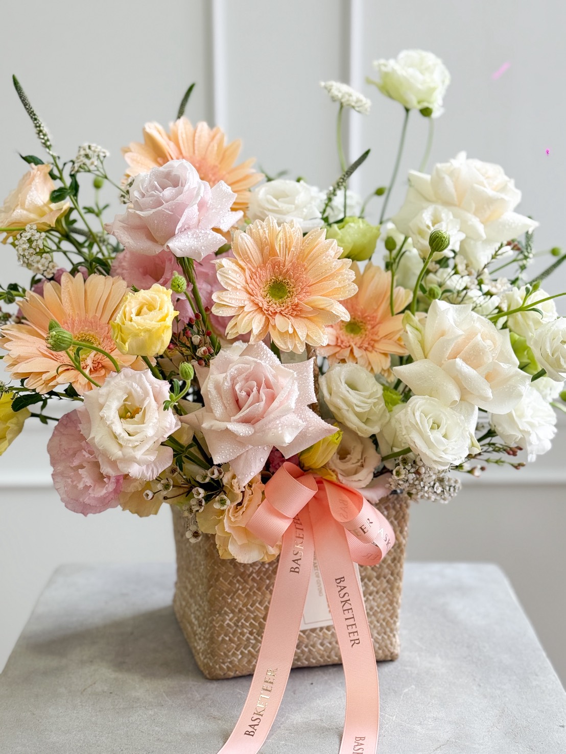 A pastel basket in a woven design featuring White O’Hara & Pink Mondial Roses, pale pink gerbera daisies, lisianthus, and lush greenery, finished with a peach 
